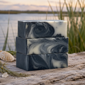 Moonlit Marsh Cleansing Bar - stacked handcrafted charcoal soap bars with black and white swirls on driftwood by a marsh